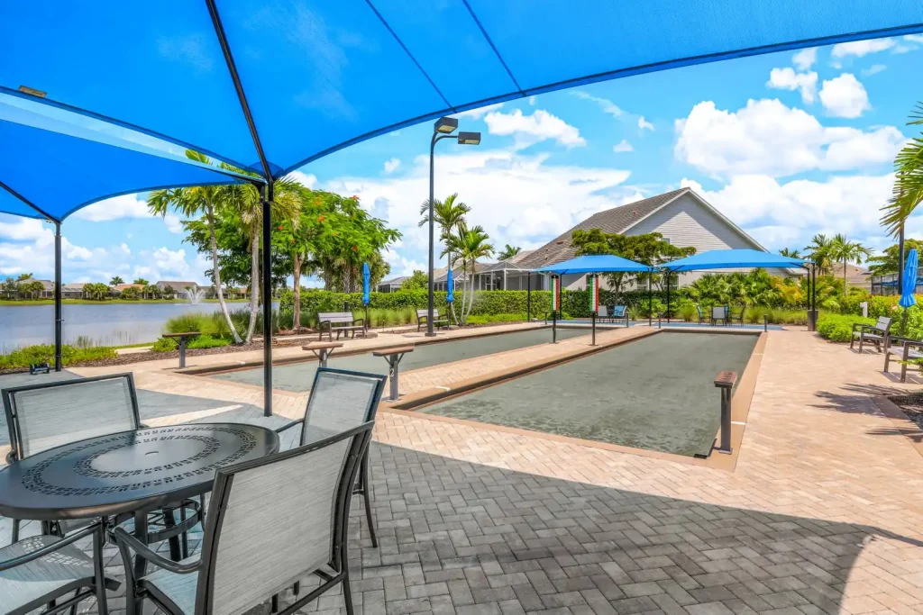 Outdoor resort shuffleboard court under blue shade sails, with tables, benches, palm trees and a lake in the background.
