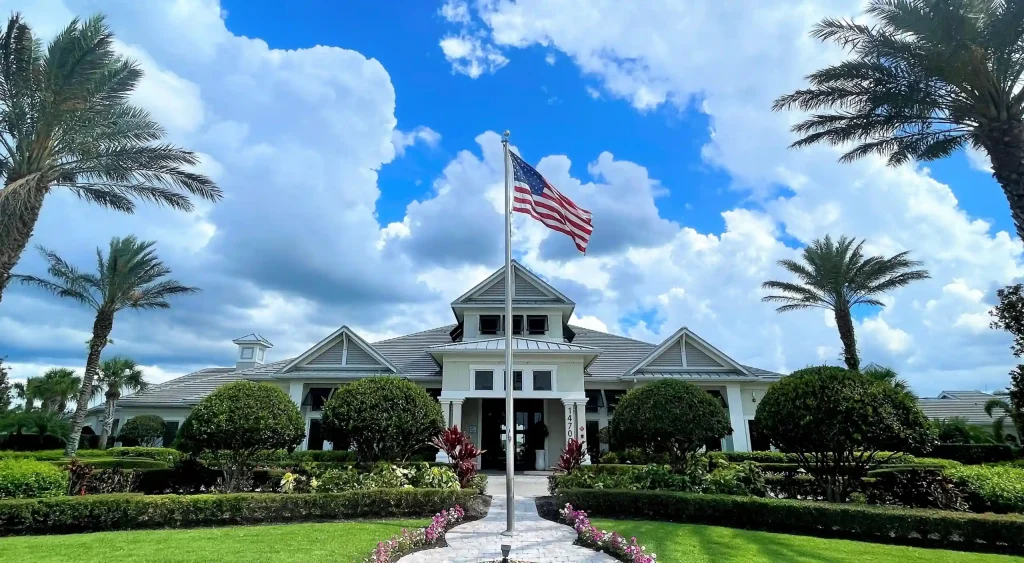 American flag waving on a flagpole in front of a white clubhouse with palm trees and neatly trimmed shrubs under a blue sky with clouds.