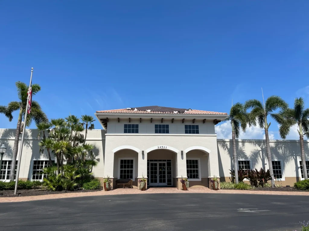 Front view of a beige building with arched entryways, palm trees, and an American flag under a blue sky.