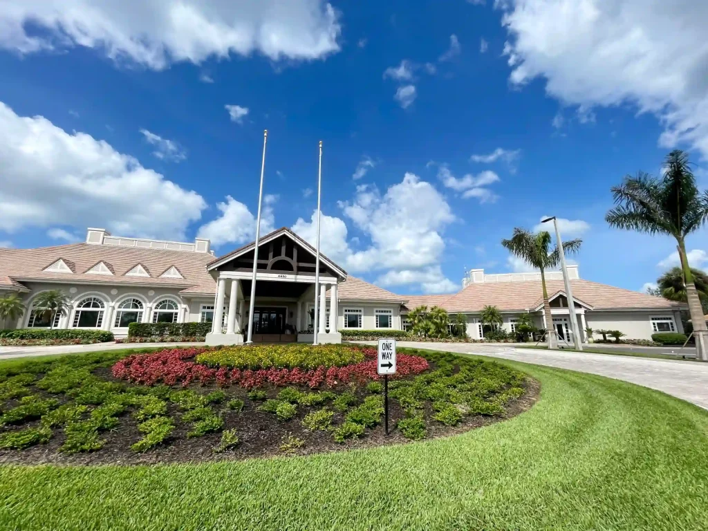 Beige campus building with arched windows, two flagpoles, and vibrant flower beds under a sunny blue sky with palm trees nearby.