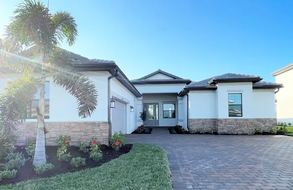 Single-story house with light siding, stone accents, and a brick-paved driveway; palm trees and shrubs flank the entrance.