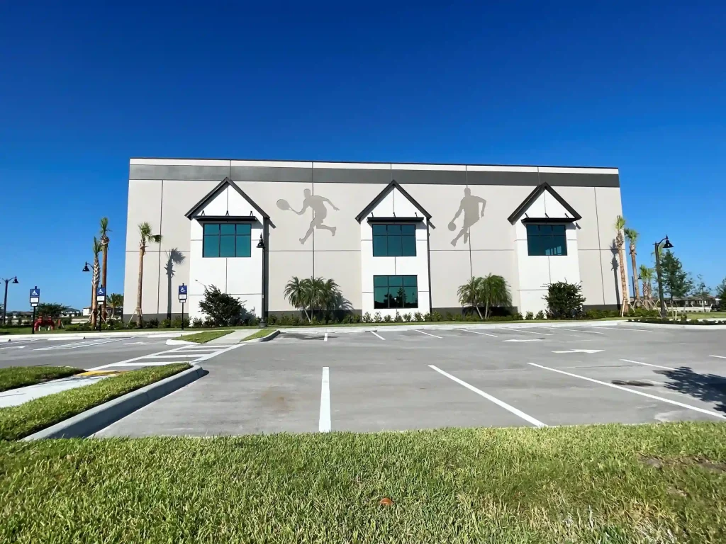 Two gray athletic silhouettes on the building’s facade, with peaked awnings, under a clear blue sky in a parking lot.