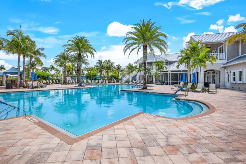 Resort pool area with palm trees, lounge chairs, and blue umbrellas under a bright sky.