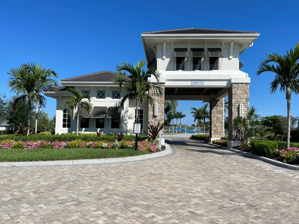White two-story building entrance with stone columns, palm trees, and a brick-paved driveway against a blue sky.